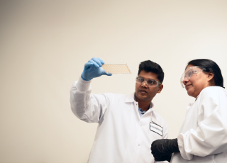 Two workers wearing PPE stand in front of a white background, looking at a piece of laboratory equipment.