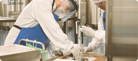 Two workers wearing white coats, blue aprons, and PPE work with equipment in a laboratory.